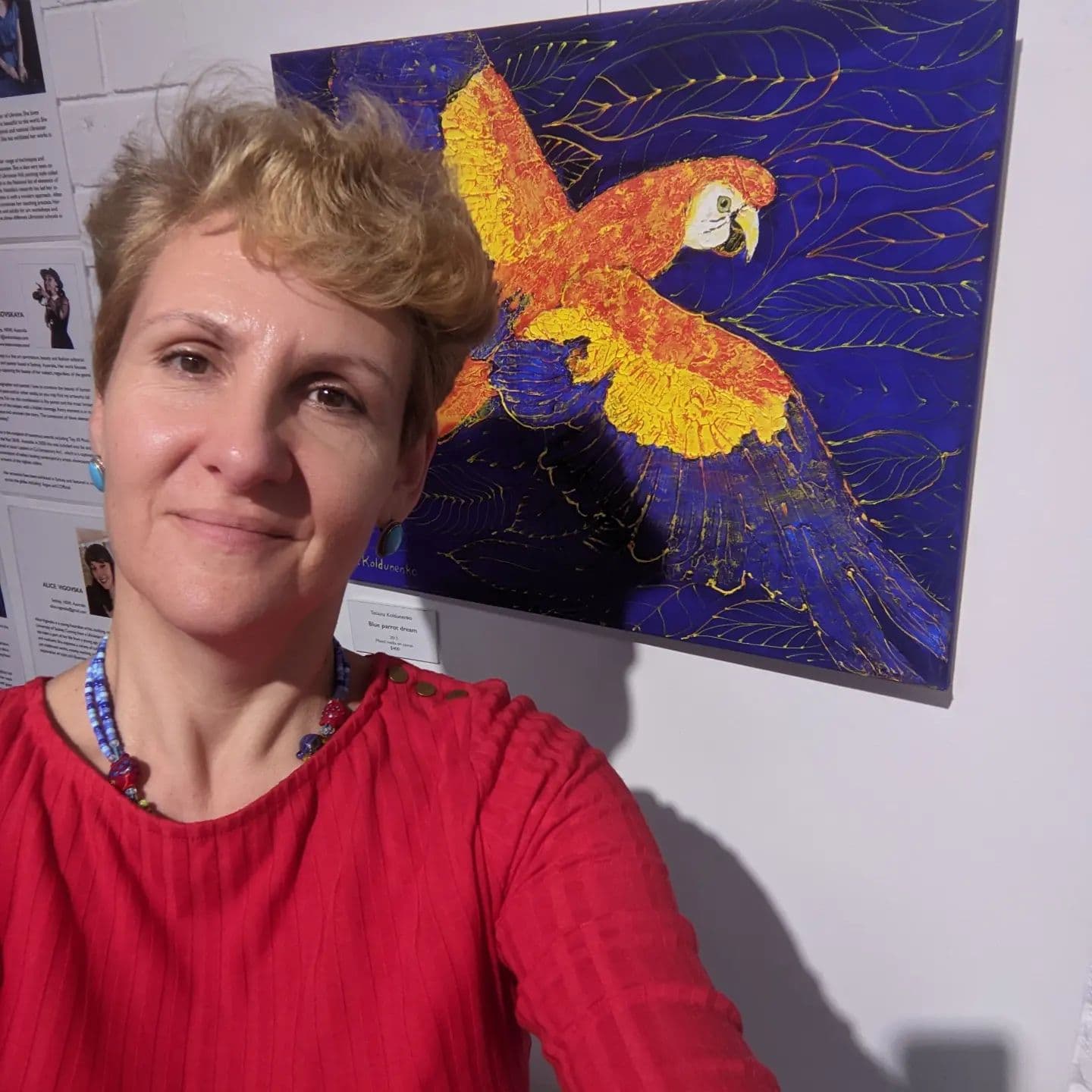Contemporary Australian artist Tetiana Koldunenko in front of her painting of bright red and yellow parrot on blue background