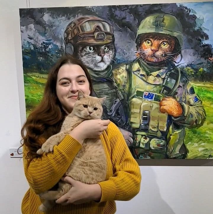 A girl holding a cat in front of a painting with 2 cats